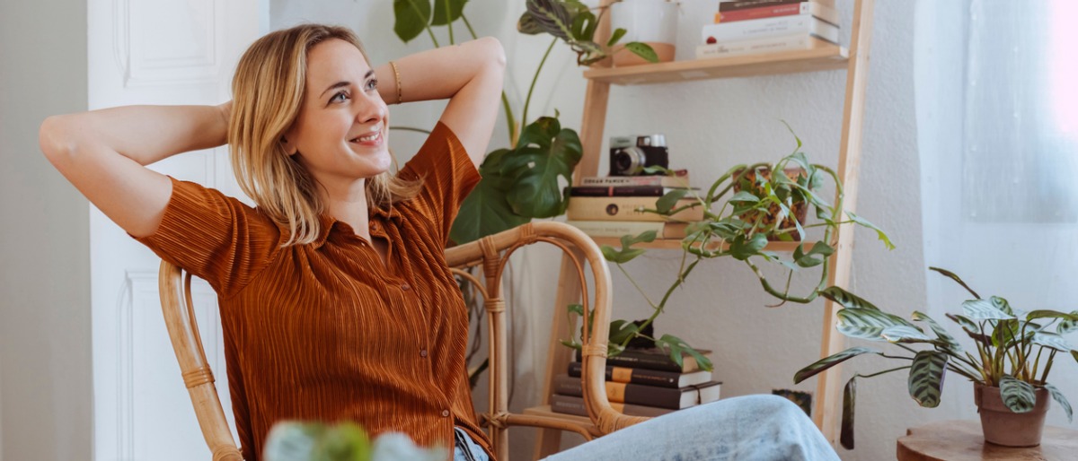 Portrait of a young happy smiling woman with raised hands behind head, sitting on the chair in her living room early in the morning and looking at the window. Copy space, interior background.